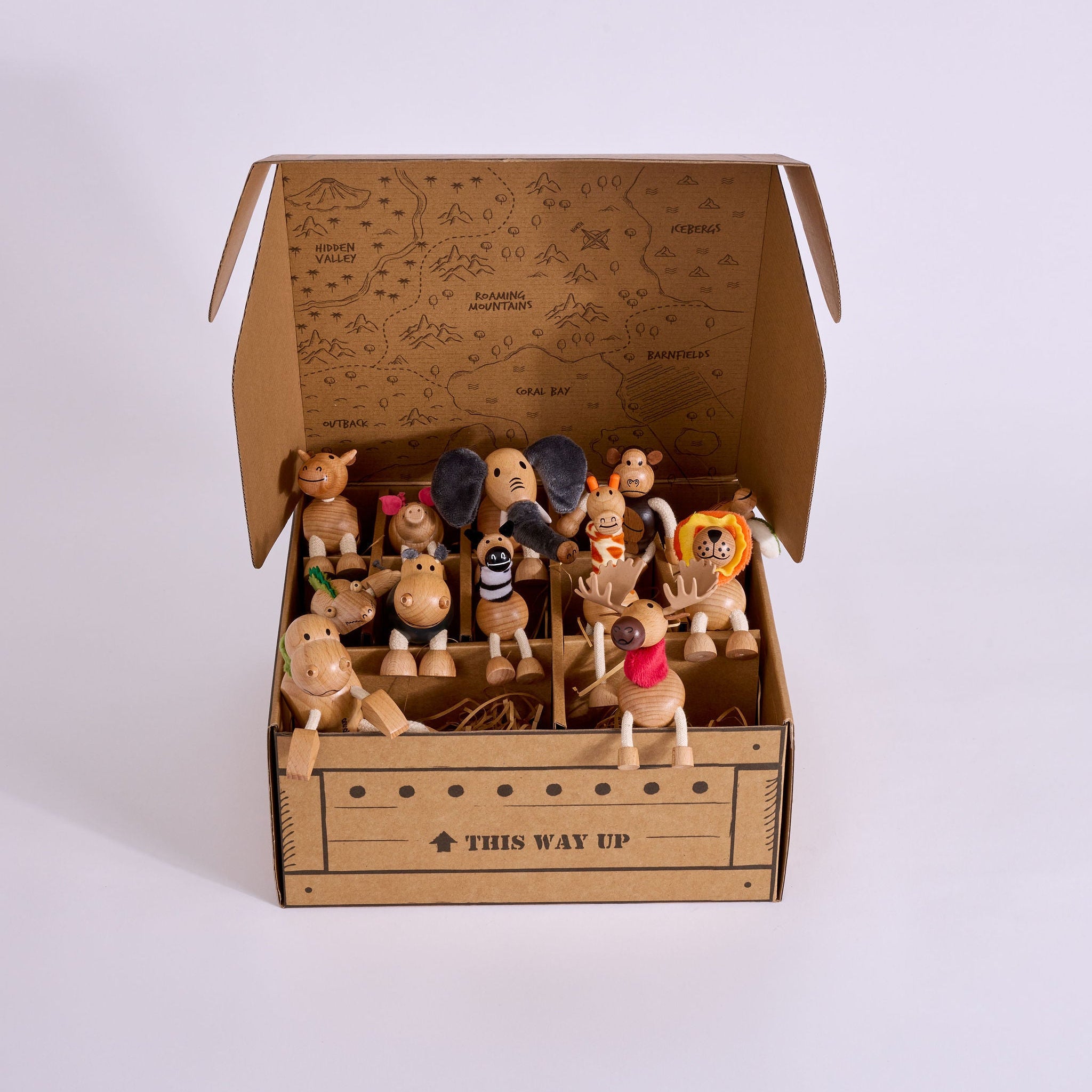 A collection of small wooden toy figurines and animal characters arranged inside an open cardboard gift box with printed map artwork on the inside lid and the text 'THIS WAY UP' on the front. The box is photographed on a clean white studio background.
