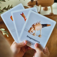 Hand holding a card with a Stegosaurus illustration on a wooden surface with a toy dinosaur in the background.