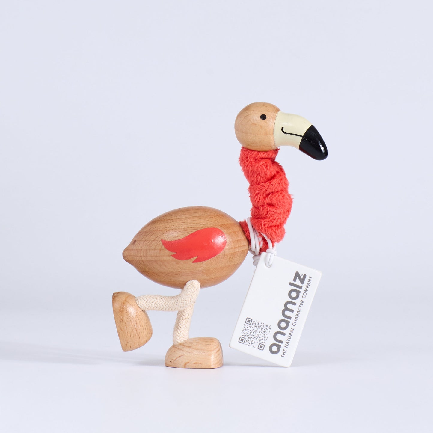 Wooden flamingo toy with a coral plush scarf and a visible brand tag on a light gray background